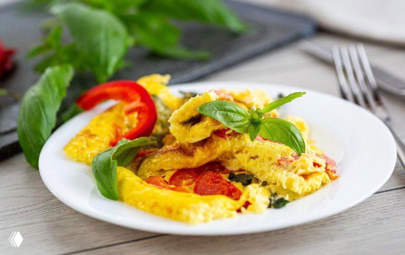 Омлет из двух яиц с овощами и свежей зеленью на тарелке, яркие кусочки перца и томата, домашняя уютная сервировка.