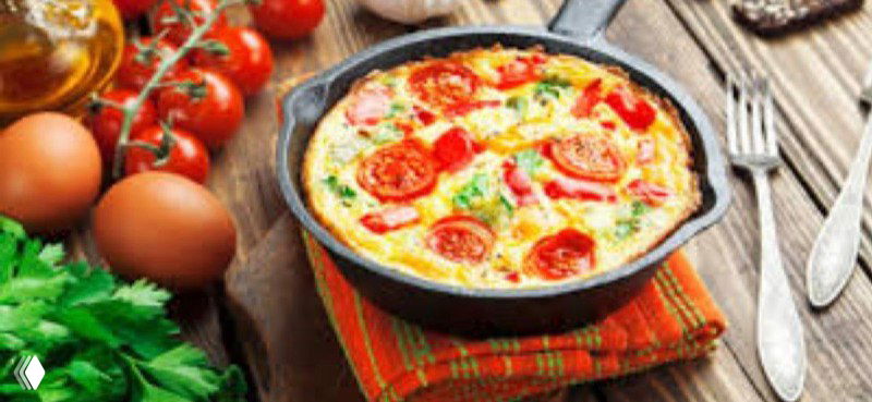 Чугунок с запечённым овощным омлетом/рагу, сверху томаты и зелень, вид сбоку на сковороду с кухонным фоном.