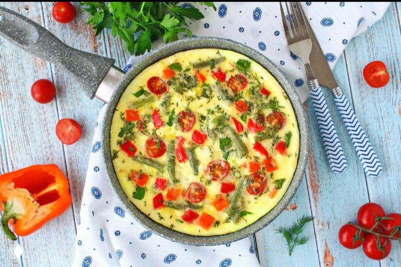 Яркий омлет с кусочками томатов и зеленью в сковороде на текстильной скатерти, летняя подача и свежая зелень рядом.