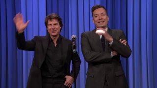 Два человека на телевизионной сцене на синем фоне: мужчина в тёмном костюме машет рукой рядом с ведущим, видна сцена и микрофон