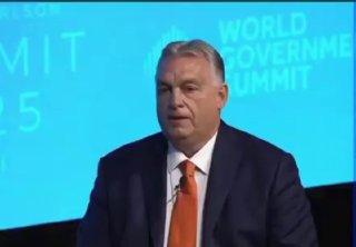 Мужчина в костюме сидит на сцене конференции, средний план, видны задник мероприятия и элементы презентации в фоне.