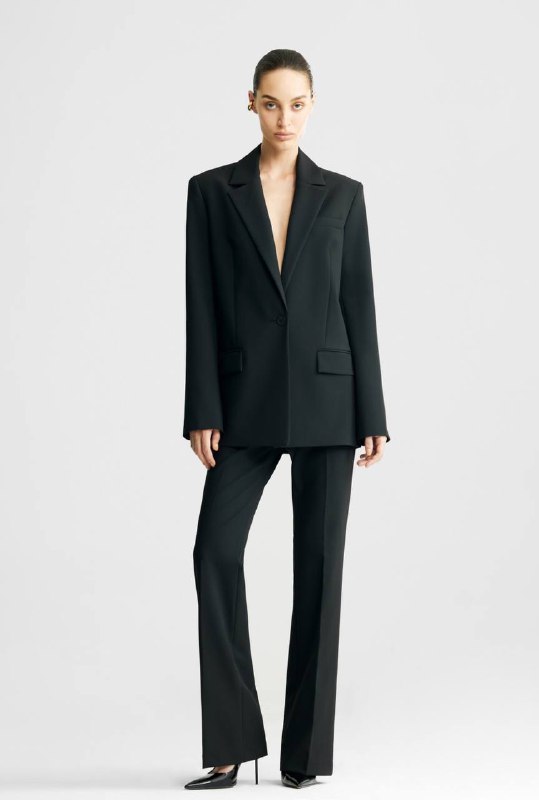 Модель в удлинённом чёрном жакете и брюках: образ в полный рост, подчёркнуты линии плеч и длина жакета, современный деловой силуэт.