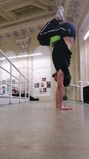 Атлет в стойке на руках без опоры, между ног зажат зелёный кубик для контроля линии тела, интерьер зала с зеркалами и перекладинами