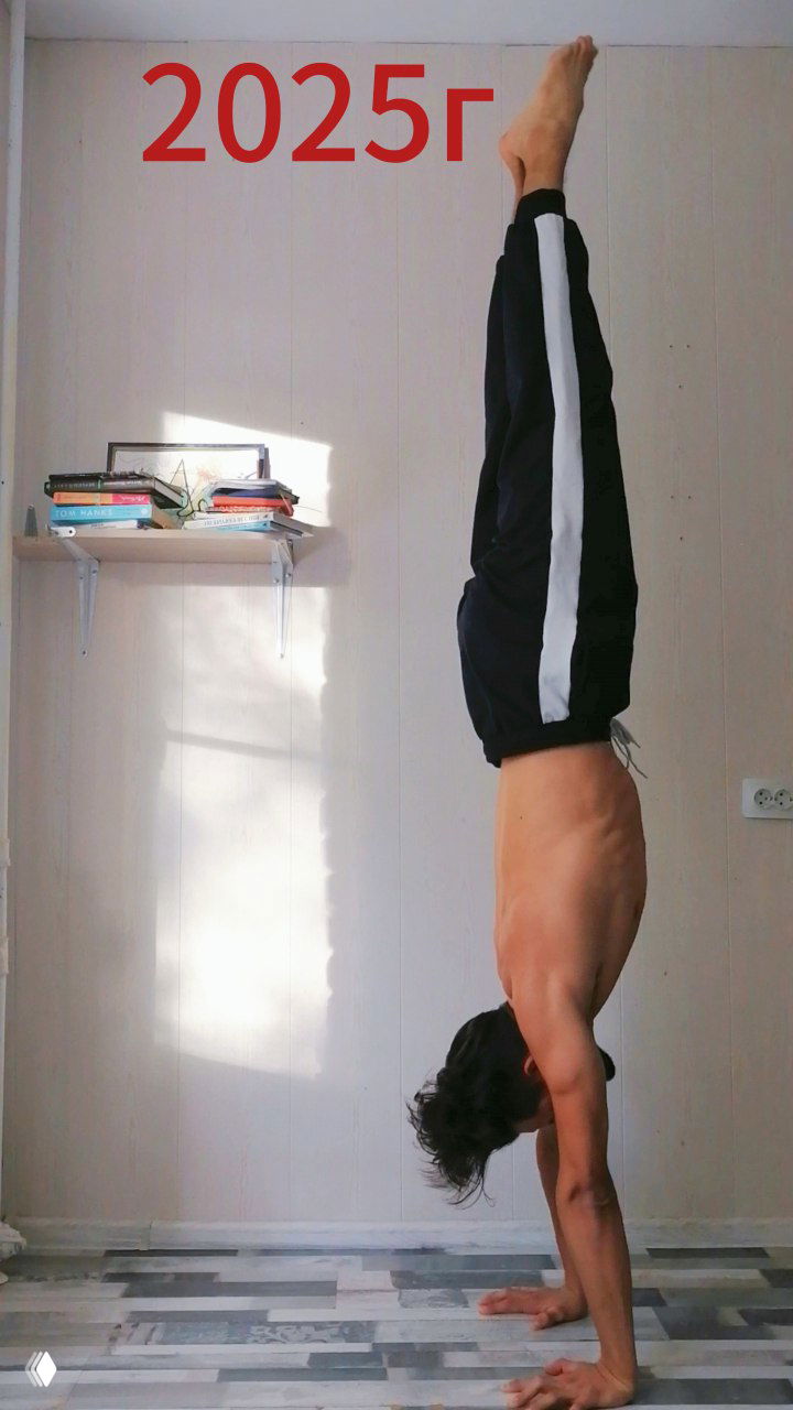 Фото 2025: человек в стойке на руках в домашнем интерьере, выровненное тело и ноги вверх, видна полка с книгами и домашняя обстановка.