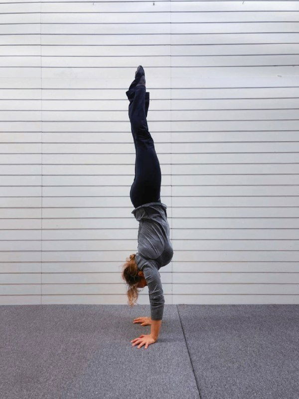 Женщина в одиночной стойке на руках у белой стеновой панели в зале — ровная вертикаль, упор на прямые плечи и контроль корпуса.