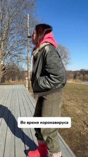 Человек в профиль на деревянной дорожке, в зелёной куртке и розовом капюшоне, солнечный день; на фото наклейка «Во время коронавируса».