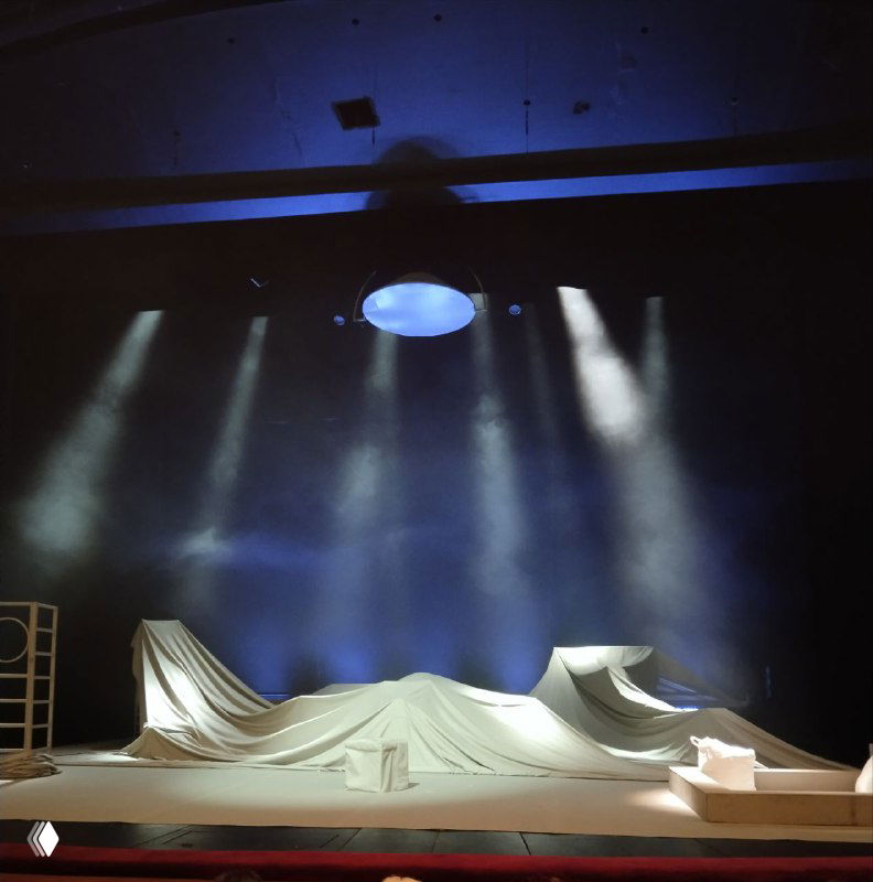 Сцена перед началом спектакля: синие софиты, свисающие элементы и накрытые драпировкой декорации — атмосферный театральный кадр.