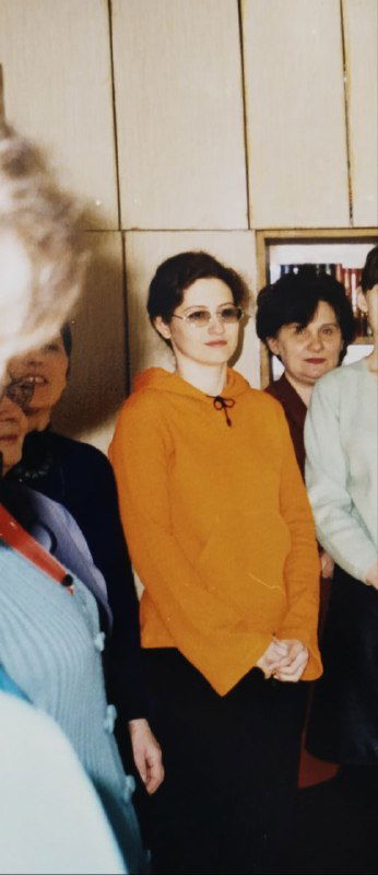 Фотография в интерьере — женщина в оранжевой кофте среди коллег у двери, начало 2000-х, официальная обстановка и серьёзный вид.