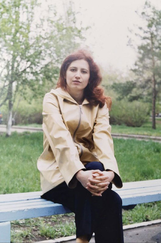 Портрет в парке: женщина в светлом плаще на скамейке, зелёный фон, снимок начала 2000-х, спокойный повседневный кадр.