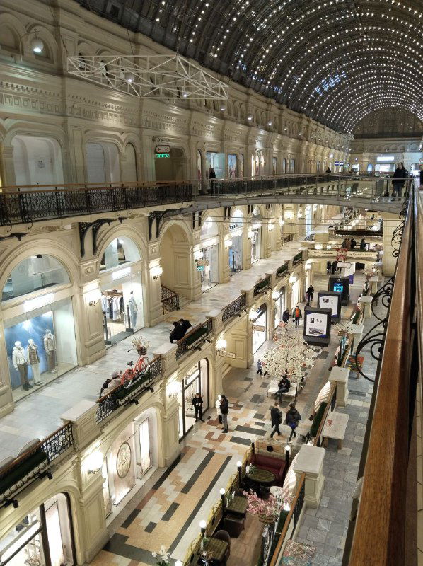 Интерьер ГУМа: двухъярусная галерея с арочными окнами, витринами магазинов и высоким стеклянным потолком, подсвеченным в вечернее время.