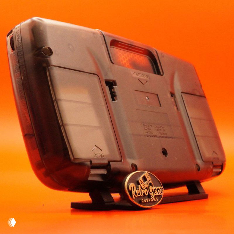 Вид сзади под углом на Game Gear: открытые отсеки батарей, подставка с логотипом RetroGear и полупрозрачный тёмный корпус на оранжевом фоне.