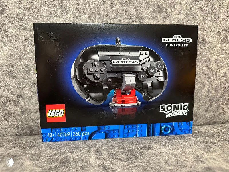 Фотография коробки набора LEGO Sonic стоящей на текстильном фоне: крупный логотип SEGA Genesis и изображение собранного контроллера в центре упаковки, тёплые оттенки освещения.