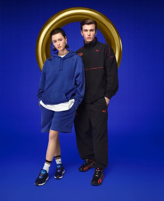 Модель и модель в синей и чёрной спортивной одежде Puma с элементами Sonic — худи и шорты в комплекте, позируют на синем фоне с реквизитом в виде кольца.