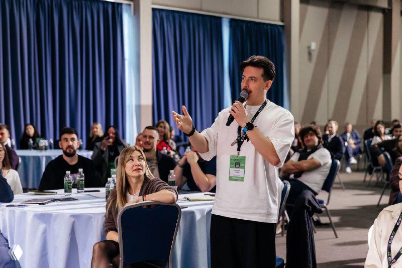 Спикер задаёт вопрос со сцены перед аудиторией: люди слушают, некоторые записывают, атмосфера деловой конференции.