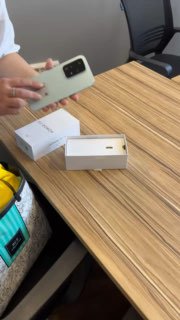 Крупный план: руки держат смартфон над распакованной коробкой на столе в офисе; видна упаковка и часть сумки внизу кадра.