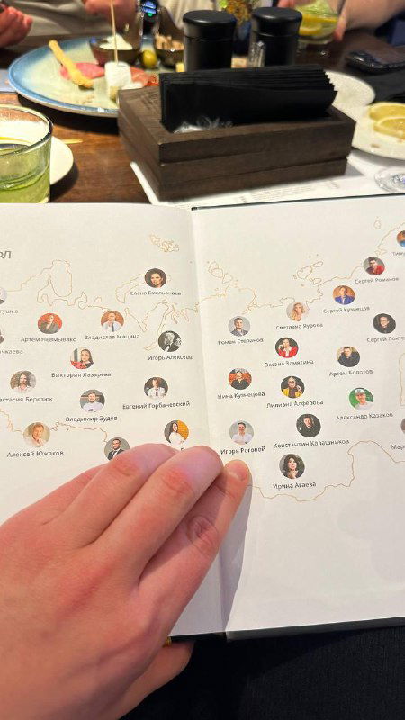 Рука удерживает раскрытый буклет или раздаточный материал с миниатюрами участников и схемой — демонстрация содержания или структуры сообщества.