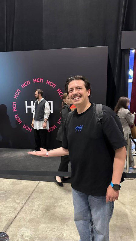 Игорь Роговой на площадке мероприятия у стенда с логотипом; в кадре он жестом показывает пространство, вокруг люди и выставочные элементы конференции.