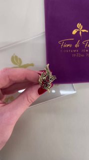 Крупный план кулона маски FUOCO из Fiore di Firenze в руках, видно оформление коробки бренда и красно‑золотые эмалевые детали изделия.