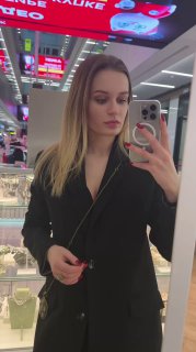 Фотография продавца/модели в зеркале в магазине — женщина в чёрном пиджаке делает селфи на фоне витрин с украшениями, интерьер ТЦ виден за спиной.