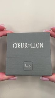 Двойной кадр серой подарочной коробки COEUR de LION в руках продавца — вид спереди с логотипом и текстурой упаковки.