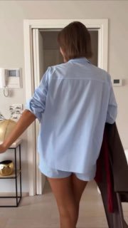 Частный кадр: женщина в голубой рубашке и коротких шортах стоит спиной в светлом интерьере, создавая уютный образ.