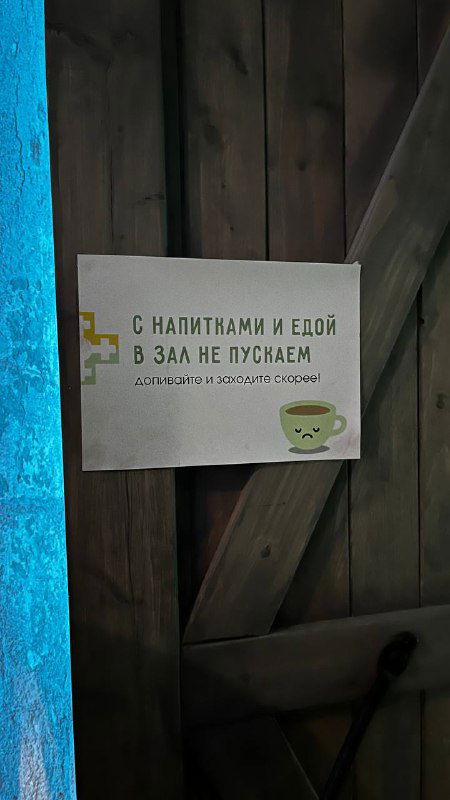 Деталь экспозиции и церковной лавки: табличка у деревянной двери с надписью о запрете с напитками и едой, часть мерча рядом.