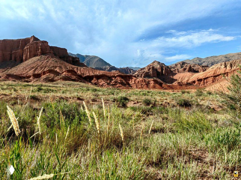 Передний план с травой и каменистым рельефом, на заднем плане — красные скальные массивы Каньона Конорчек и ясное небо.