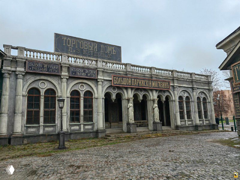 Неоклассический торговый дом с вывесками и аркадой на пустой брусчатой площади — декорация, создающая атмосферу начала XX века.