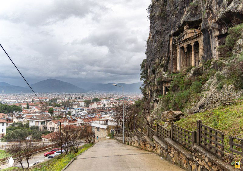 Дорога вдоль скалы и гробница Аминтоса высеченная в скале: деревянный забор, каменные ступени и вид на город внизу создают атмосферу археологического места.