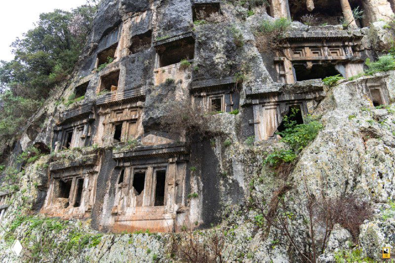 Фасад ликийских гробниц в скале: ряд резных ниш и входов, множественные захоронения, их каменная архитектура и естественное разрушение временем хорошо видны в кадре.