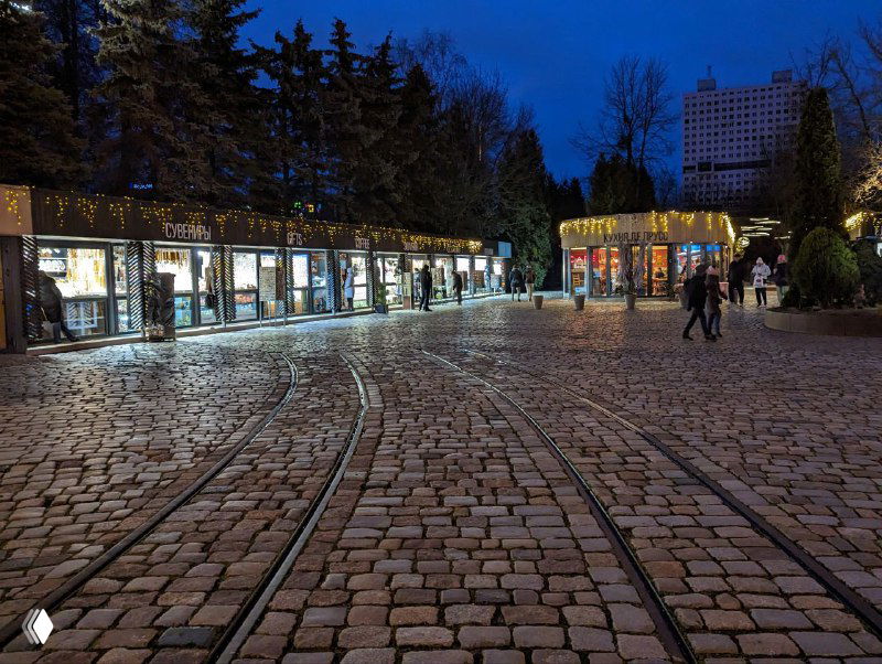 Пешеходная площадь у Рыбной деревни в Калининграде с брусчаткой и трамвайными рельсами, вечерняя сцена с витринами и редкими прохожими.