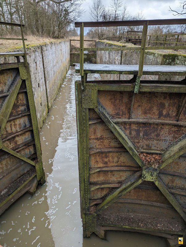 Вид сверху на железные перекладины и узкий водный коридор шлюза №2; мутная вода, мох и облупившаяся краска на металле.