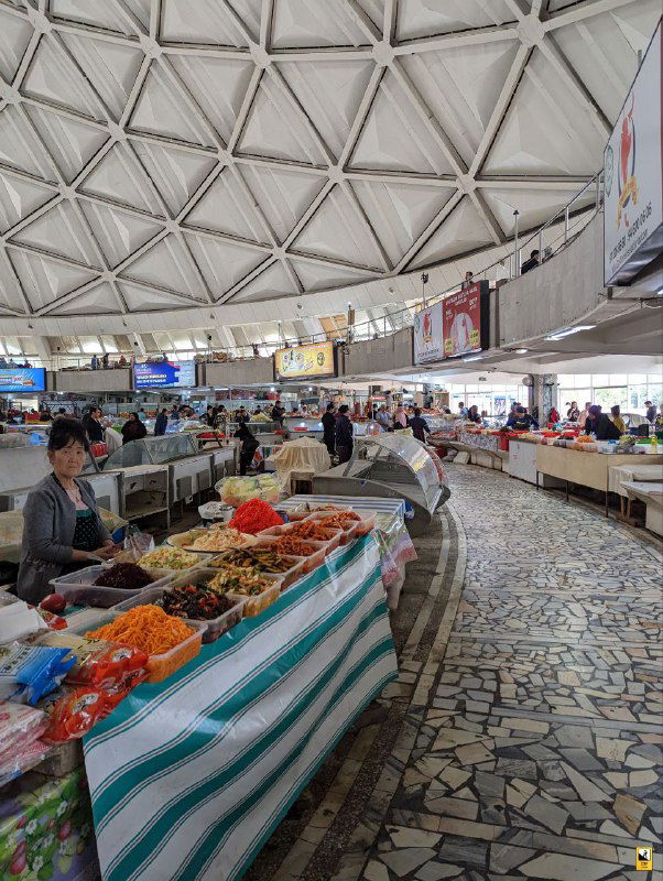 Круглая крытая часть рынка с торговыми прилавками внутри: соленья, сухофрукты и продукты аккуратно выложены, торговцы за прилавками и мозаичный пол заметны в кадре.