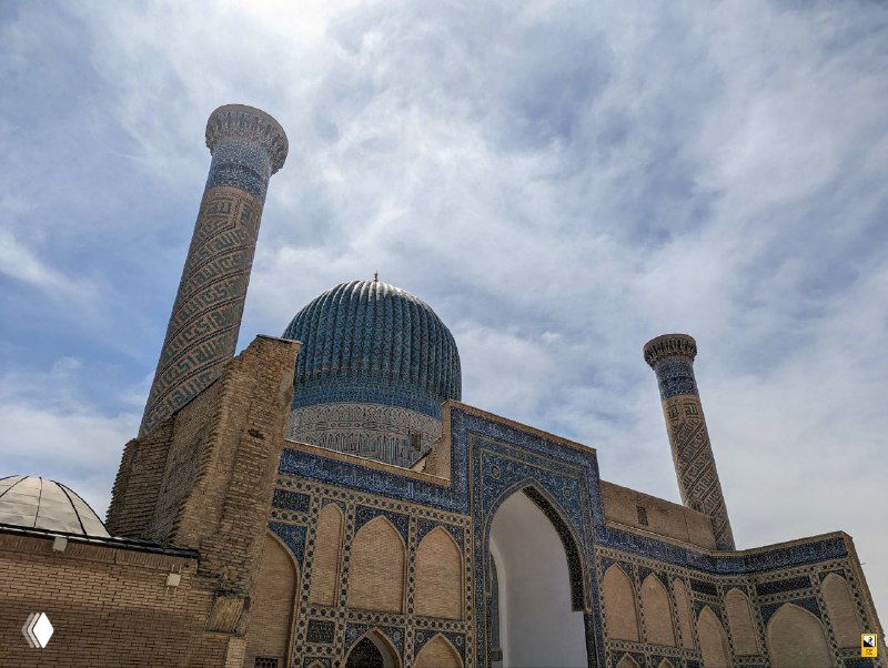 Дневной ракурс на главный купол и минареты комплекса Гур-Эмир: бирюзовый купол, узкие цилиндрические минареты и фасад из кирпича и плитки.