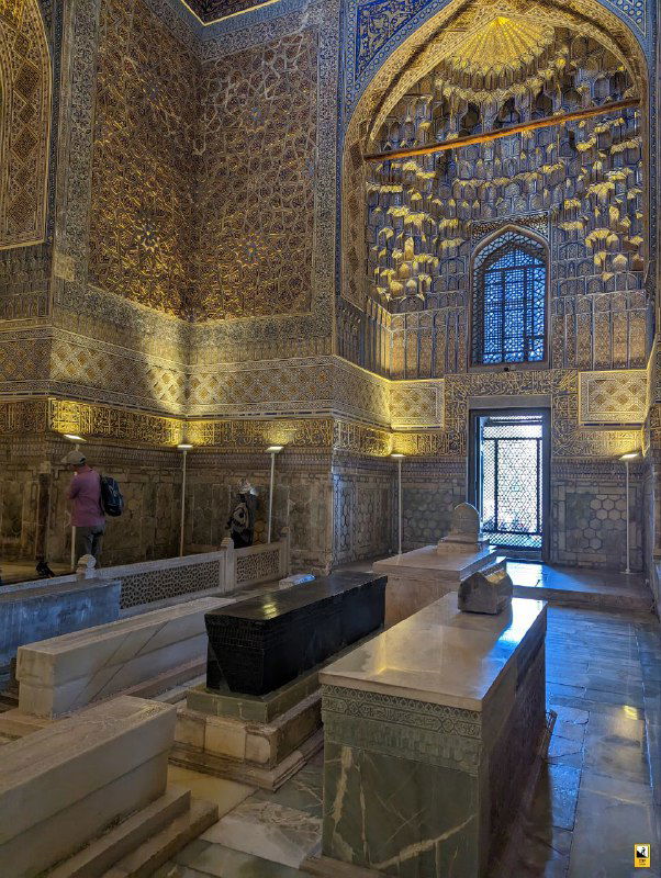 Зал с надгробиями внутри Гур-Эмир: каменные саркофагоподобные надгробия на фоне мозаичного декора стен и окон, посетители в кадре.