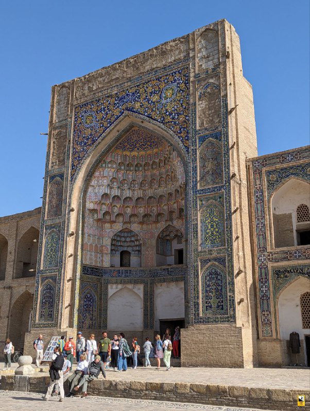 Высокий портал медресе с плиточной мозаикой в Бухаре; у основания арки собрались туристы, снимающие фасад и архитектуру.