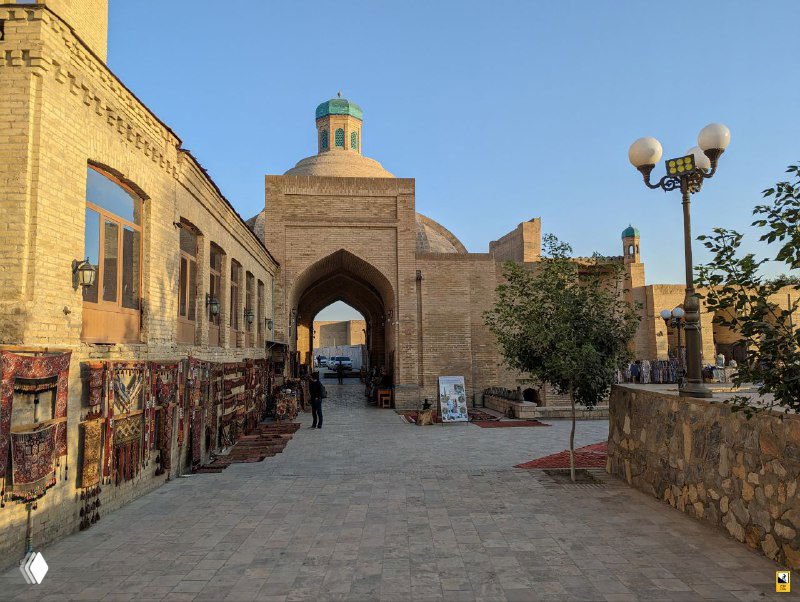 Арочный вход к торговой площади в Бухаре, по бокам висят ковры и сувениры; несколько посетителей прогуливаются по вымощенной улице.