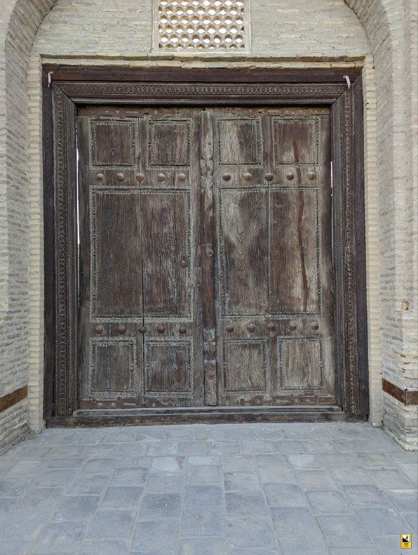 Старая массивная деревянная дверь на мощёной улице Бухары; текстура дерева и каменная кладка подчёркивают вековую атмосферу места.