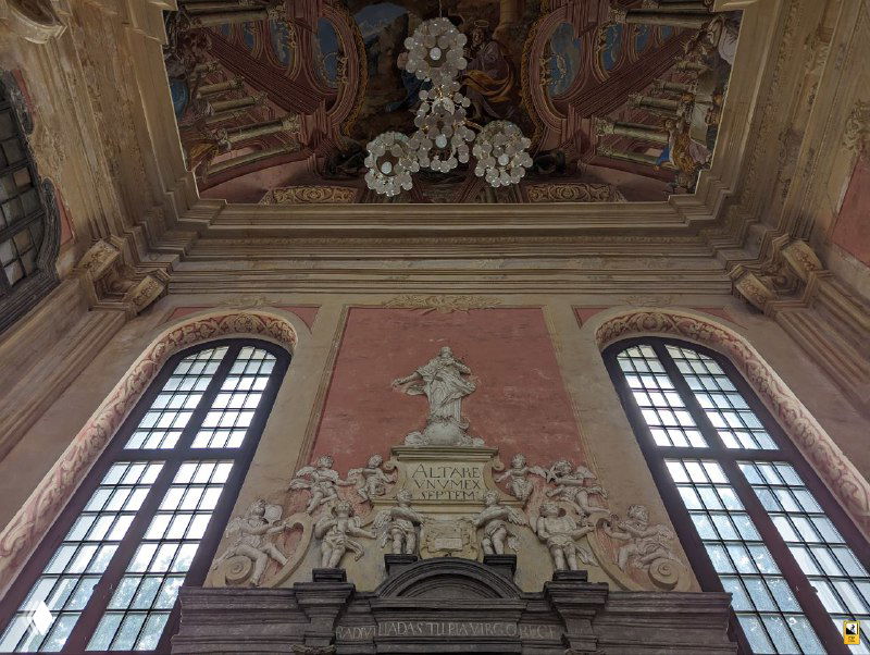 Вид на часть внутренней стены с лепным декором и высоким окном: сочетание барочного орнамента и световых акцентов внутри помещения.