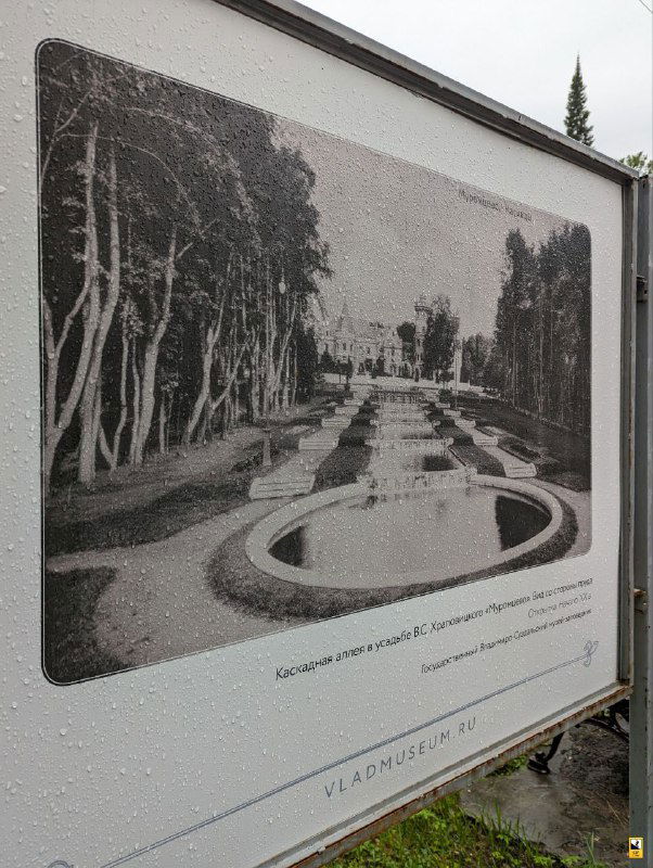 Информационный щит с архивной фотографией каскада прудов усадьбы Муромцево и сопровождающей подписью, фото сделано в дождливую погоду.