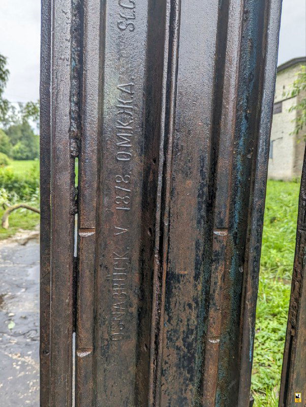 Крупный план металлической рельсовой детали, ранее принадлежавшей железной дороге усадьбы, сейчас используемой как ограждение с видимыми надписями.