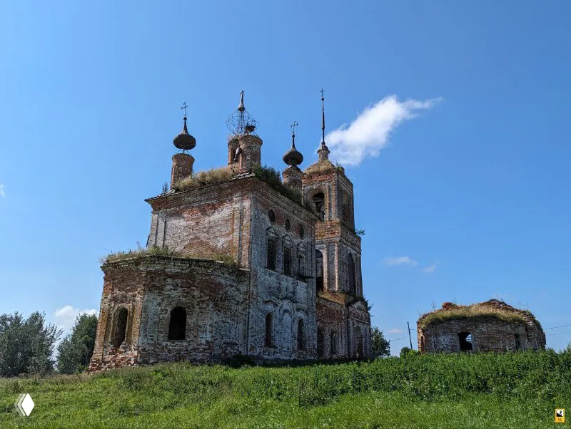 Общий боковой вид церкви Флора и Лавра в Киболе со стороны опушки поля; руины колокольни и основного объёма здания под синим небом.