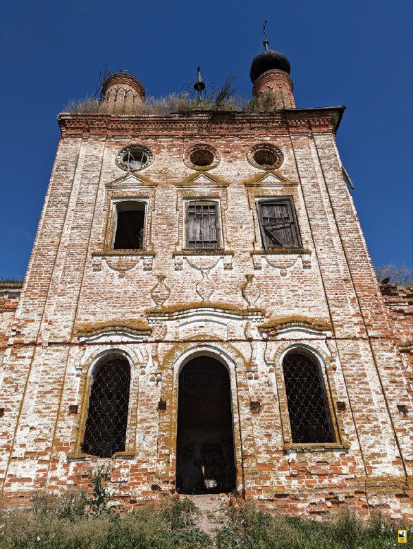 Фасад и главный вход церкви Флора и Лавра с тремя арочными порталами и обрушившейся отделкой; заросли и следы разрушения у основания.