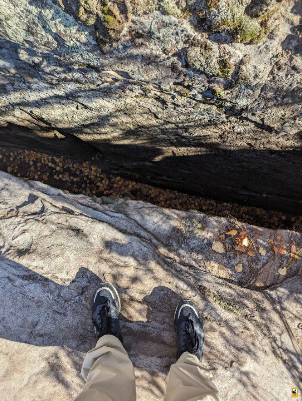 Каменная поверхность вблизи с видимыми трещинами, текстурой песчаника и ботинками автора на краю скалы, листья осени вокруг.