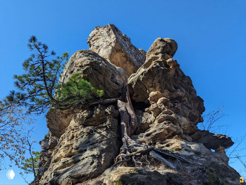 Каменный столб и причудливые скальные выступы в портретном кадре, осенние цвета вокруг и ясное небо наверху.