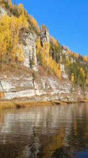Крупный фрагмент скалы и берега Усьвы в осенней окраске; вид сбоку на каменные образования, листва и спокойную воду под голубым небом.