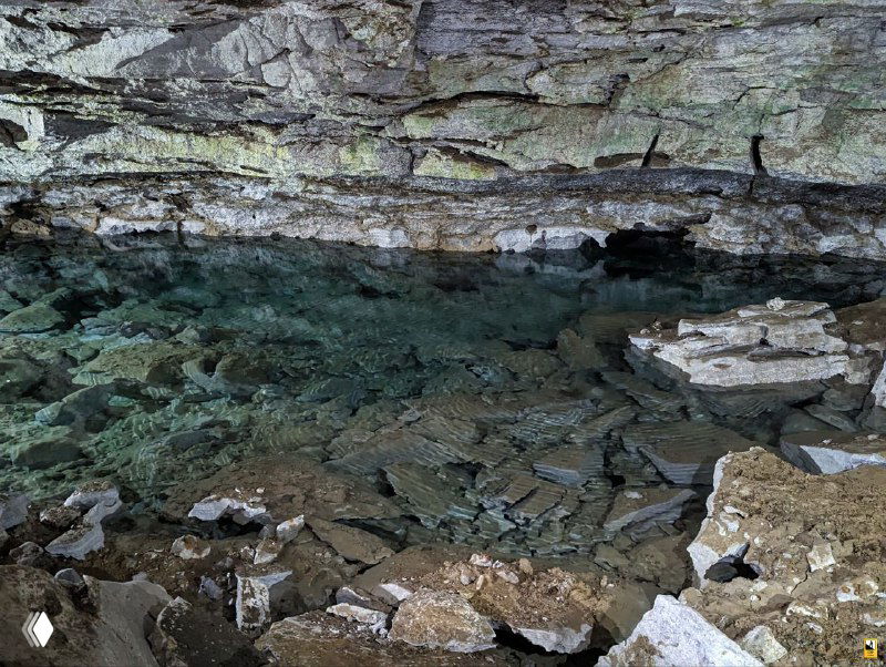 Прозрачная подземная вода в карстовом пруду пещеры, ровное зеркало воды и обломки пород по берегам; подчёркнута глубина и структура дна, фото из экскурсии.