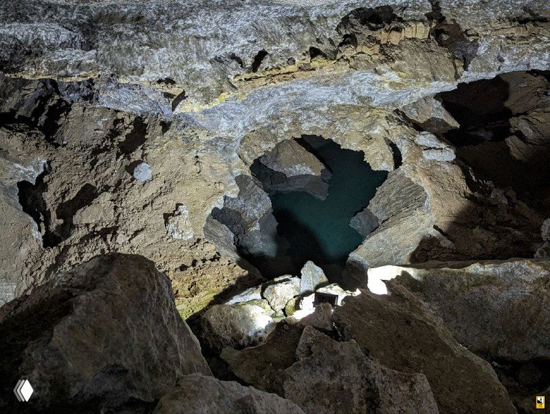 Карстовая воронка с тёмной водой внизу, вид сверху: освещение акцентирует трещины, слои пород и контрасты каменной структуры внутри Кунгурской пещеры.