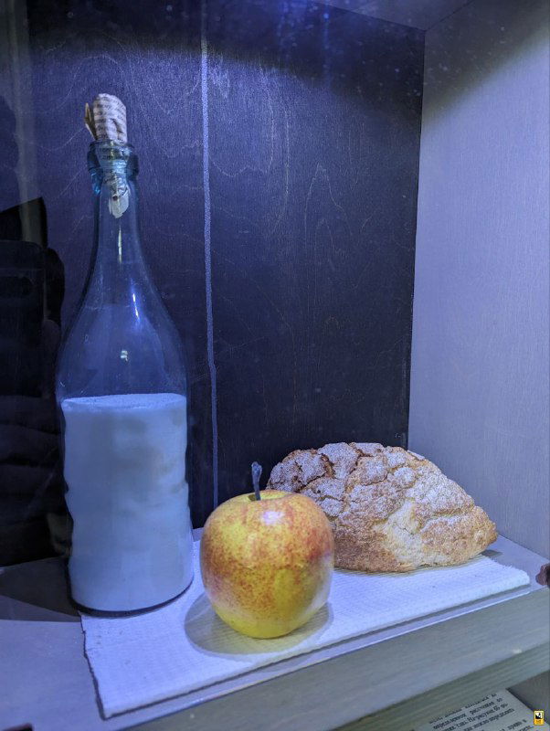 Реплика завтрака ученика в экспозиции музея: бутылка молока, хлеб и яблоко, выставленные на ткани как образ повседневного школьного питания того времени.