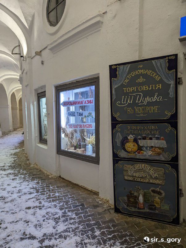 Улица с витриной магазина и табличкой в историческом проходе Красных рядов в Костроме, заснеженная тропинка рядом с витриной.